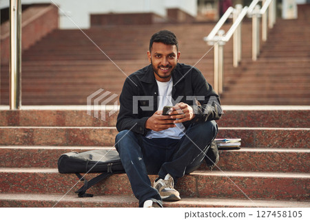Handsome Indian student is outdoors sitting on the stairs with smartphone 127458105