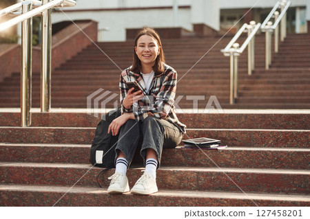 Smartphone in hands. Young female student is sitting on the stairs of university 127458201