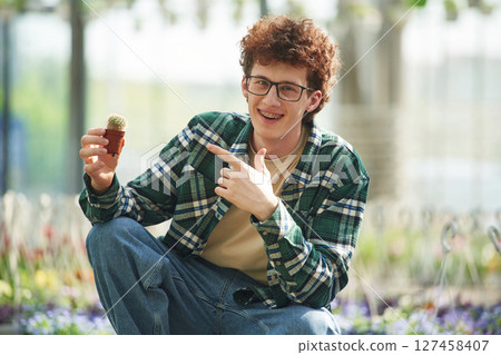 Sitting and holding the little cactus. Young man with curly hair and in glasses is in greenhouse Sitting and holding the little cactus. Young man with curly hair and in glasses is in greenhouse 127458407