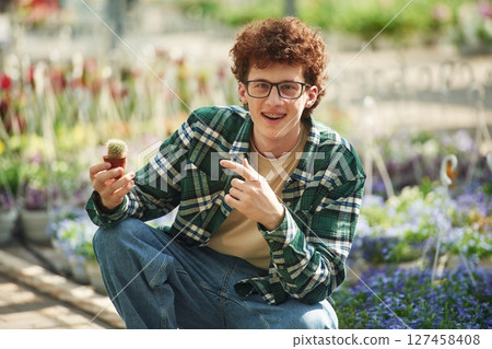 Sitting and holding the little cactus. Young man with curly hair and in glasses is in greenhouse Sitting and holding the little cactus. Young man with curly hair and in glasses is in greenhouse 127458408