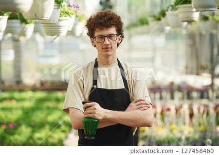 Arms crossed. With watering bottle. Young man with curly hair and in glasses is in greenhouse 127458460