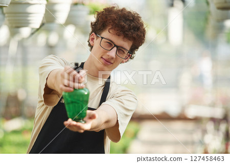 Front view, standing with watering bottle. Young man with curly hair and in glasses is in greenhouse 127458463