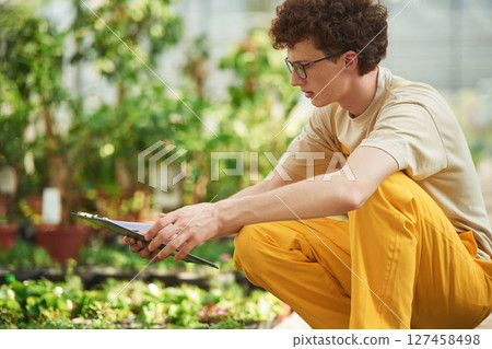 Quality control, with notepad. Young man with curly hair and in glasses is in greenhouse 127458498