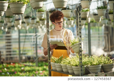 Notepad in hands. Young man with curly hair and in glasses is in greenhouse Notepad in hands. Young man with curly hair and in glasses is in greenhouse 127458567