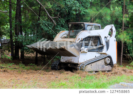 Compact loader working on landscape clearing in wooded area during overcast leveling landscaping Compact loader working on landscape clearing in wooded area during overcast leveling landscaping 127458865
