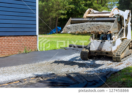 Gravel being spread by machinery along residential foundation in landscaping project during ground leveling Gravel being spread by machinery along residential foundation in landscaping project during ground leveling 127458866