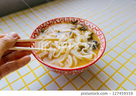 person is eating noodles with chopsticks. colorful bright red plate of ramen sits on yellow table.  127459359