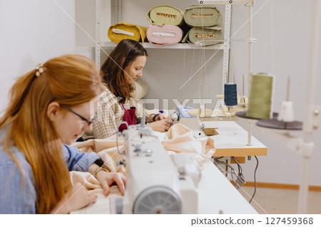 Two female seamstresses in the workshop at the factory are working at tables on sewing machines. Two female seamstresses in the workshop at the factory are working at tables on sewing machines. 127459368