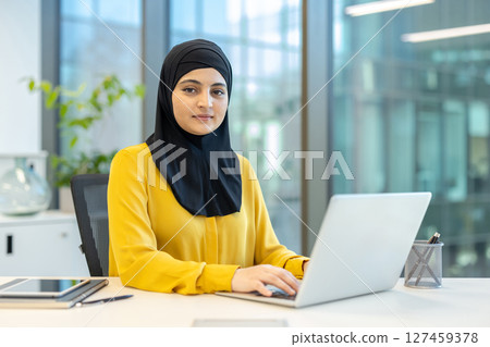 A woman wearing a hijab sits at a desk working on a laptop in a modern office setting. She is focused and appears to be working professionally. 127459378