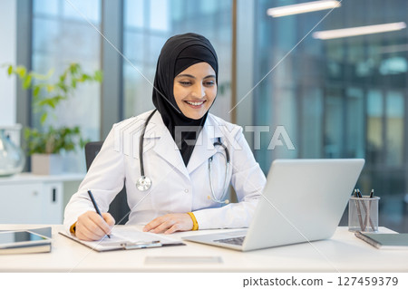 A smiling female doctor, wearing a hijab, works at a desk with a laptop, taking notes, in a modern office setting. A smiling female doctor, wearing a hijab, works at a desk with a laptop, taking notes, in a modern office setting. 127459379