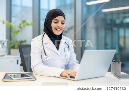 An arab female doctor, wearing a hijab, smiles while working on a laptop at her desk. The image is taken in a modern office setting. An arab female doctor, wearing a hijab, smiles while working on a laptop at her desk. The image is taken in a modern office setting. 127459380