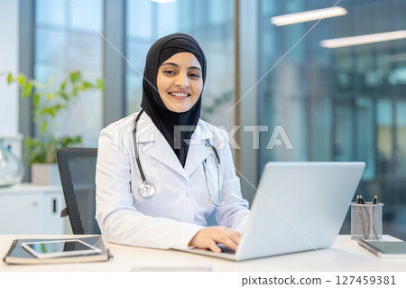 A smiling female doctor wearing a hijab sits at a desk, working on a laptop in a modern office setting, focusing on her work with a friendly demeanor. A smiling female doctor wearing a hijab sits at a desk, working on a laptop in a modern office setting, focusing on her work with a friendly demeanor. 127459381