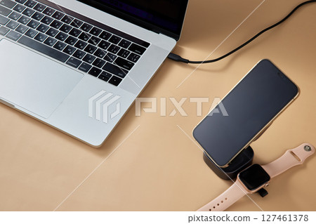 Laptop with smart watch and smartphone charging on wireless charger on beige table, Modern workplace device 127461378
