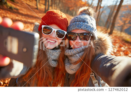 Two young women taking a selfie in a vibrant autumn forest with colorful leaves surrounding them 127462012