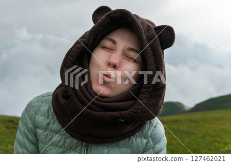 Cute young woman traveler in down sweater and fleece hoodie with bear ears blowing kiss to camera with closed eyes against natural evening landscape 127462021