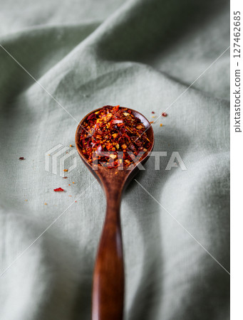 Wooden spoon filled with crushed red chili flakes on green fabric background 127462685