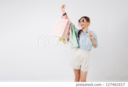 Happy young Asian woman shopping on Black Friday raising her hand with shopping bags, cheerful and excited holding colorful bags after shopping, isolated studio white background 127462817
