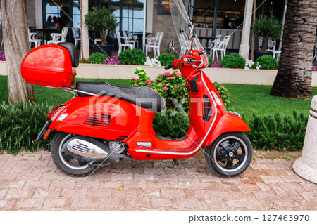 Red scooter parked on a city street near cafe. Urban mobility, personal transport, and summer commuting. Red scooter parked on a city street near cafe. Urban mobility, personal transport, and summer commuting. 127463970