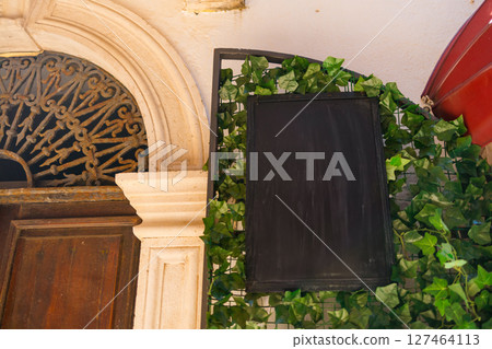 Empty chalkboard sign framed with green ivy beside vintage doorway. Hospitality decor, rustic signage, and the aesthetic charm of local cafes and restaurants. 127464113
