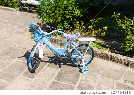 Blue kids bicycle with training wheels on sunny pavement. Childhood fun, outdoor learning, and the joy of early cycling experiences in a quiet garden path. Blue kids bicycle with training wheels on sunny pavement. Childhood fun, outdoor learning, and the joy of early cycling experiences in a quiet garden path. 127464116