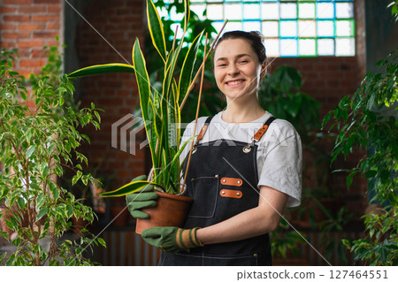 Running of own business. Young woman florist holding plant in pot wearing apron in botanical store. Happy small business owner working at flower shop smiling surrounded by plants. Small business 127464551