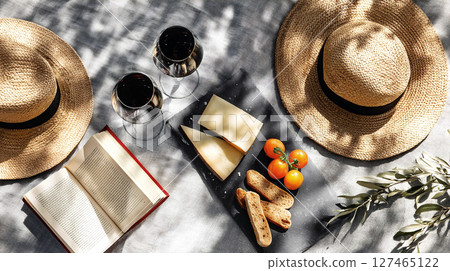 Two straw hats, glasses of red wine, cheese, and snacks on a picnic table, with an open book and fresh tomatoes, creating a relaxing outdoor dining atmosphere Two straw hats, glasses of red wine, cheese, and snacks on a picnic table, with an open book and fresh tomatoes, creating a relaxing outdoor dining atmosphere 127465122