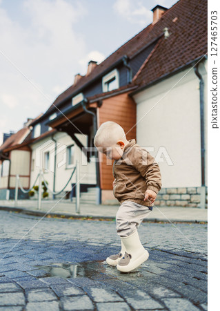 A 2-year-old boy in a light jacket and rubber boots runs through puddles after the rain. A boy in boots stands in a puddle. The child rejoices in the rain and runs along the wet street. A 2-year-old boy in a light jacket and rubber boots runs through puddles after the rain. A boy in boots stands in a puddle. The child rejoices in the rain and runs along the wet street. 127465703