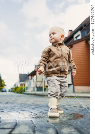 A 2-year-old boy in a light jacket and rubber boots runs through puddles after the rain. A boy in boots stands in a puddle. The child rejoices in the rain and runs along the wet street. A 2-year-old boy in a light jacket and rubber boots runs through puddles after the rain. A boy in boots stands in a puddle. The child rejoices in the rain and runs along the wet street. 127465704