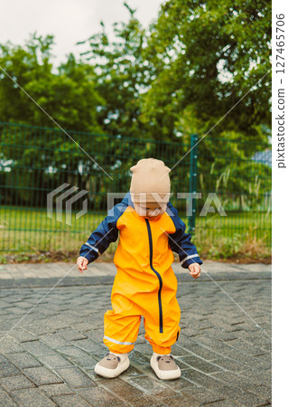 Smiling 2 year old boy in mudproof overalls and boots runs after the rain. Toddler in bright raincoat and boots runs through puddles. Child in water-repellent overalls on a walk on a cloudy day. Smiling 2 year old boy in mudproof overalls and boots runs after the rain. Toddler in bright raincoat and boots runs through puddles. Child in water-repellent overalls on a walk on a cloudy day. 127465706