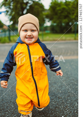 Cheerful boy 2 years old in yellow waterproof overalls walking along asphalt road on rainy day. Stylish boy in yellow waterproof jacket and rubber boots playing outdoors. Cheerful boy 2 years old in yellow waterproof overalls walking along asphalt road on rainy day. Stylish boy in yellow waterproof jacket and rubber boots playing outdoors. 127465707