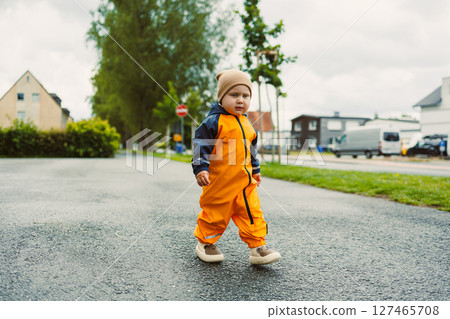 Happy 2 year old toddler in orange raincoat playing in puddle on wet road. Child on rainy day outdoors in waterproof clothes and rubber boots. Joy of childhood and active recreation. Happy 2 year old toddler in orange raincoat playing in puddle on wet road. Child on rainy day outdoors in waterproof clothes and rubber boots. Joy of childhood and active recreation. 127465708