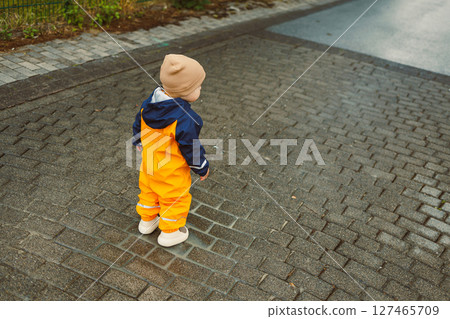 Cheerful boy 2 years old in yellow waterproof overalls walking along asphalt road on rainy day. Stylish boy in yellow waterproof jacket and rubber boots playing outdoors. Cheerful boy 2 years old in yellow waterproof overalls walking along asphalt road on rainy day. Stylish boy in yellow waterproof jacket and rubber boots playing outdoors. 127465709