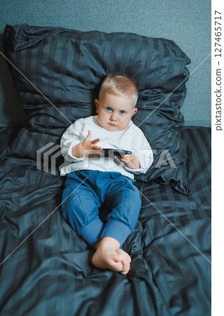 Boy holds phone in hands. Handsome toddler boy in white t-shirt and jeans sits on bed and watches cartoons on phone. 2 year old boy plays with mobile gadget. Modern digitalization of kids. Boy holds phone in hands. Handsome toddler boy in white t-shirt and jeans sits on bed and watches cartoons on phone. 2 year old boy plays with mobile gadget. Modern digitalization of kids. 127465717
