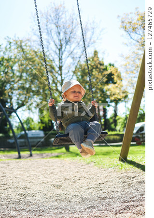 Cute 2 year old boy in a Panama hat on his head and a light jacket swings on a swing. Children's swing on a playground in the park. Toddler rides on a swing. Stylish children's clothes. Cute 2 year old boy in a Panama hat on his head and a light jacket swings on a swing. Children's swing on a playground in the park. Toddler rides on a swing. Stylish children's clothes. 127465729