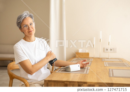 Senior woman checking blood pressure with monitor at home, sitting calmly at wooden table indoors 127471715
