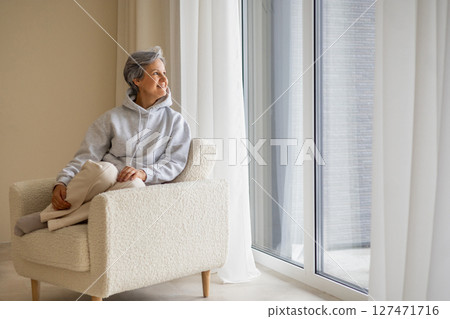Peaceful mature woman in hoodie sitting in cozy chair looking out large window at home interior 127471716