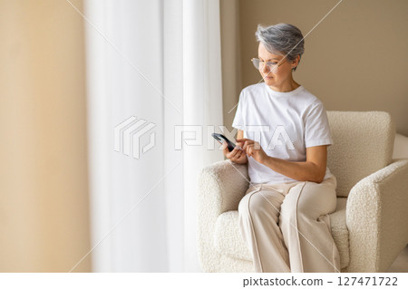 Senior woman using smartphone at home sitting in soft armchair near window in modern bright room Senior woman using smartphone at home sitting in soft armchair near window in modern bright room 127471722