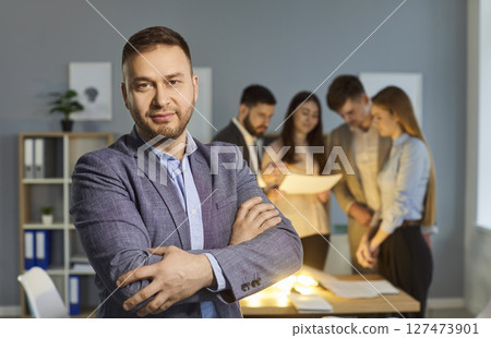Confident businessman posing with arms crossed while colleagues working on new ideas in background. Confident businessman posing with arms crossed while colleagues working on new ideas in background. 127473901