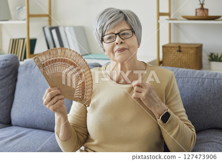 Overheated senior woman sitting on sofa and waving wooden hand fan while trying to cool down at home 127473906
