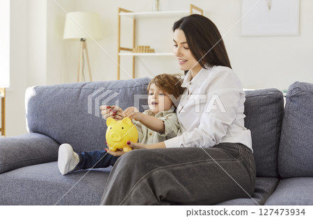 Mother and son, nanny woman sitting together on couch, interacting with yellow piggy bank, learning 127473934