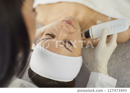 Beautician performing ultrasonic facial cleansing with ultrasonic device on woman during treatment. 127473943