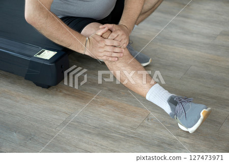 Individual stretching leg while sitting on treadmill, focusing on knee pain relief during workout session. Close up Individual stretching leg while sitting on treadmill, focusing on knee pain relief during workout session. Close up 127473971