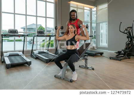 A woman perform a shoulder press with dumbbell while seated on a workout bench. Her trainer guide her, providing support and encouragement in a well-lit gym setting. A woman perform a shoulder press with dumbbell while seated on a workout bench. Her trainer guide her, providing support and encouragement in a well-lit gym setting. 127473972