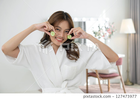 Young asian woman dressed in comfortable robe holding cucumber slice, smile while sitting in front of vanity mirror table at dressing room. Young asian woman dressed in comfortable robe holding cucumber slice, smile while sitting in front of vanity mirror table at dressing room. 127473977