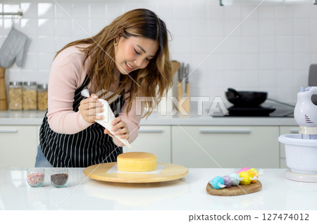 Young woman in an apron using a piping bag, pipe the icing onto the cake, creating a decorative pattern on the top of a round, light-colored cake that's placed on a wooden turntable or cake stand. Young woman in an apron using a piping bag, pipe the icing onto the cake, creating a decorative pattern on the top of a round, light-colored cake that's placed on a wooden turntable or cake stand. 127474012
