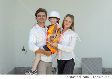 A family smile while standing on a construction site. The father hold their child in a safety vest and helmet while the mother stand beside them, both wearing casual clothing. 127474056