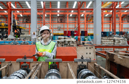 A woman in hardhat and safety vest operates industrial equipment inside a busy factory setting, skillfully managing the machinery. 127474156