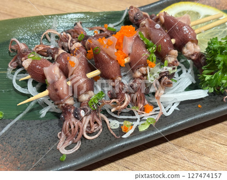 Hotaru ika sashimi skewer are elegantly presented on a black plate, accompanied by fresh vegetable and vibrant garnish, showcasing a culinary delight. 127474157