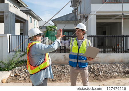 Two construction worker engage in a friendly exchange at a building site, discussing plan and progress under clear sky. The atmosphere is cooperative and productive. 127474216