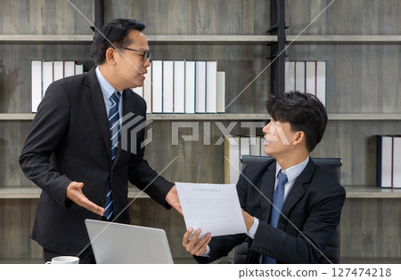 Two professional men engage in a discussion in a modern office. One man passionately speak and the other listens attentively while holding a contract document, seated at a desk. 127474218
